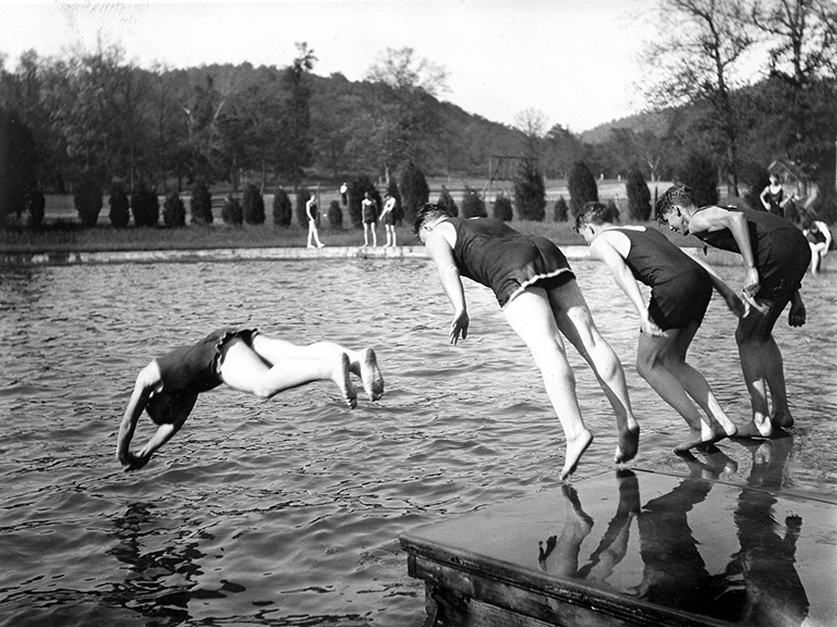 Four boys in swimming costumes with their backs to the camera are in the process of jumping off a platform into a circular pool.  About 5 other boys are in the distance, sitting and standing outside the pool looking on.