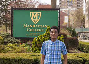 student in blue shirt in front of green manhattan college sign outside campus