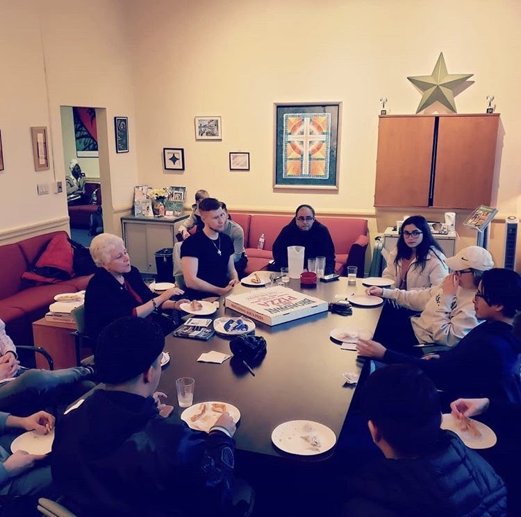 Photograph of students and faculty at a table enjoying pizza and a conversation.