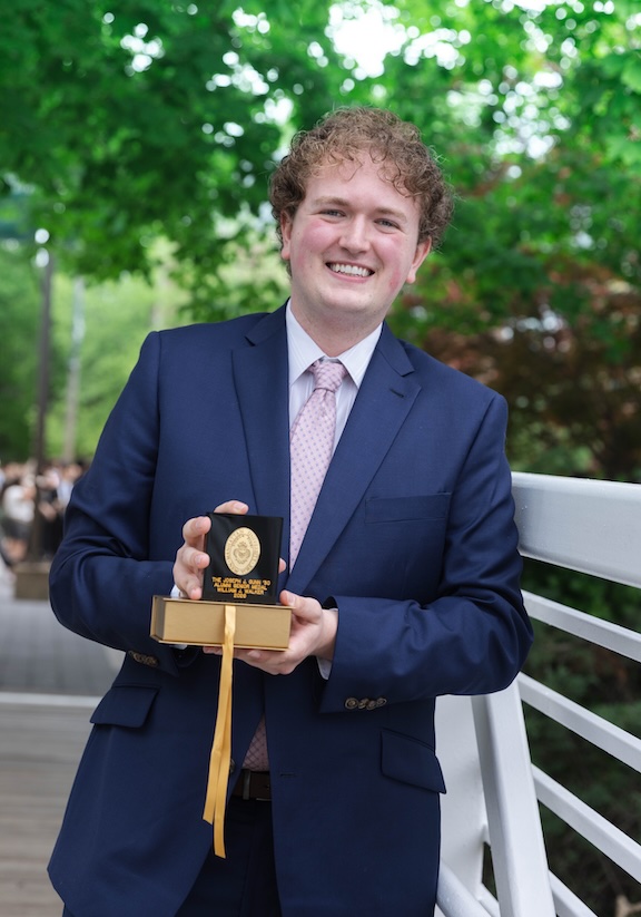 William Walker ’26 poses outdoors holding the Joseph J. Gunn ’30 Alumni Medal awarded by Manhattan University for leadership and service to the University community.